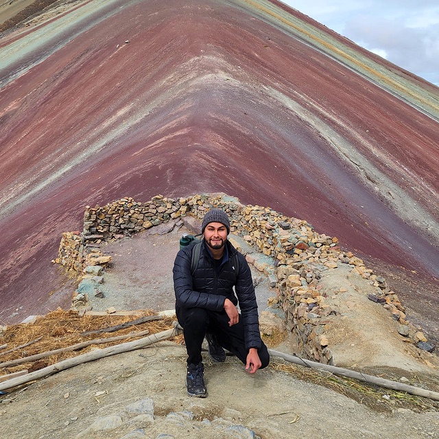 Andy kneeling on a trail in front of a rainbow mountain striped red, grey and green