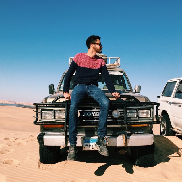 Andy sitting on the hood of an old Toyota 4x4 on desert sand under a clear blue sky